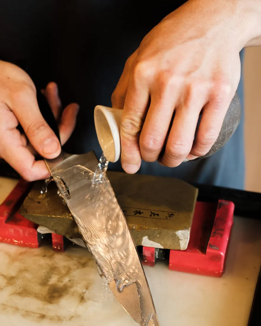 Manos afilando el filo de un cuchillo sobre una piedra de agua, con el ángulo mantenido de forma constante